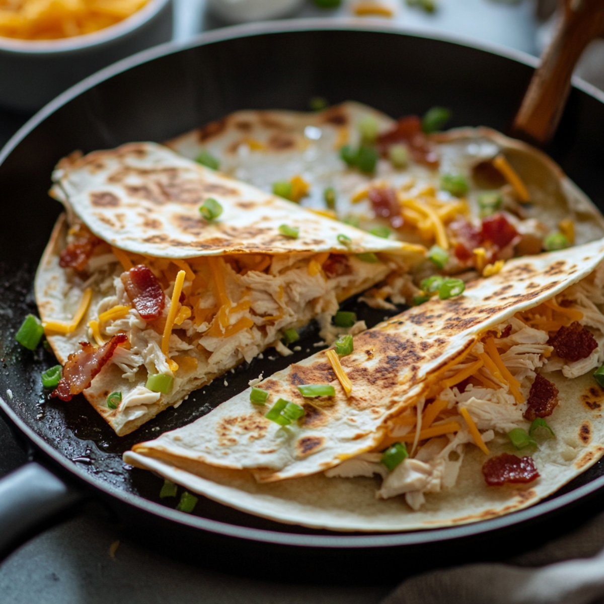 Homemade chicken bacon ranch quesadillas cooking in a skillet, filled with chicken, bacon, cheese, and green onions, with golden toasted tortillas and a wooden spatula nearby.