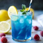 A chilled glass of homemade blue raspberry lemonade with ice, a lemon slice, fresh mint, and a striped straw, surrounded by lemons and raspberries on a marble surface.