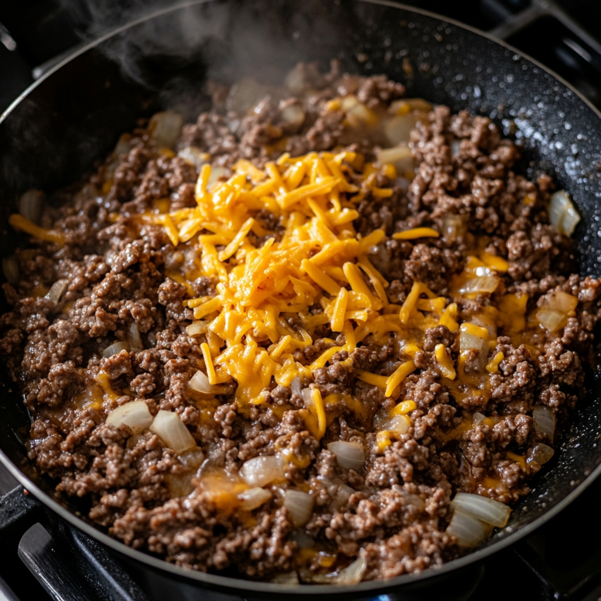 Close-up of a skillet with ground beef and diced onions, topped with shredded cheddar cheese starting to melt. Steam rising, warm natural lighting, homemade and freshly cooked feel.