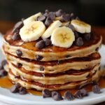 A stack of fluffy chocolate chip pancakes topped with banana slices and extra chocolate chips, drizzled with maple syrup on a white plate. Cozy and homemade.