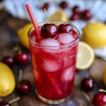 A glass of homemade cherry lemonade with ice, topped with fresh cherries and a red straw, surrounded by lemons and cherries on a wooden surface.
