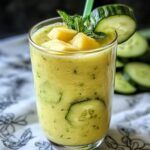 Homemade pineapple cucumber smoothie in a clear glass, topped with fresh pineapple chunks, mint, and a cucumber slice. Visible cucumber pieces inside add a fresh, natural look.