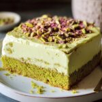 A slice of pistachio milk cake with a moist pistachio sponge base and creamy white frosting, topped with crushed pistachios and rose petals. The cake looks homemade with soft lighting and a simple plate.