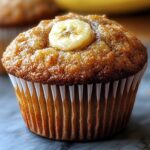 Close-up of a golden-brown sourdough banana muffins in a white paper liner, topped with a banana slice. Soft lighting, real and homemade look.