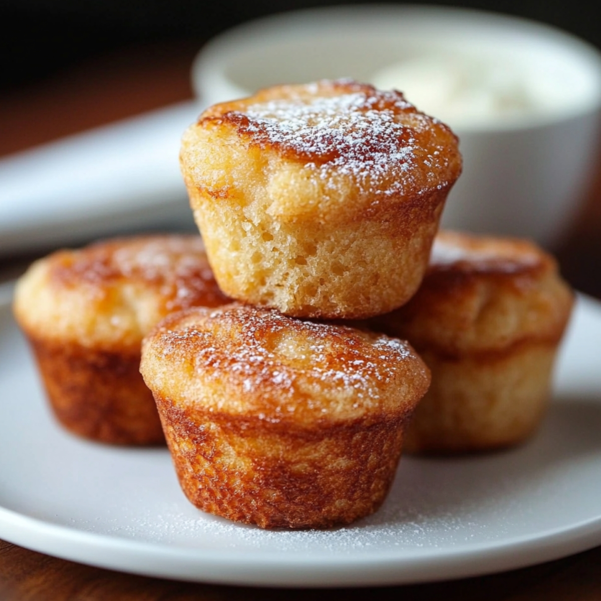 Golden brown homemade French toast muffins stacked on a white plate, lightly dusted with powdered sugar, with a blurred bowl of cream in the background.