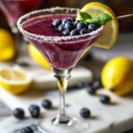 Homemade blueberry lemon drop martini in a sugar-rimmed glass, garnished with blueberries, lemon slice, and mint, with fresh fruit on marble background.