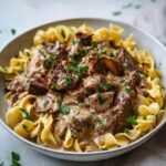 A hearty bowl of homemade beef stroganoff served over a bed of tender egg noodles. The dish features generous chunks of seared beef in a rich, creamy mushroom sauce, speckled with flecks of red pepper flakes and finely chopped fresh parsley. The sauce clings deliciously to the noodles and beef, giving a glossy, velvety finish. The noodles are cooked to a soft, slightly curled texture and sit nestled beneath the savory mixture. The dish is presented in a wide, off-white ceramic bowl on a light-colored surface, garnished with scattered parsley leaves for a fresh, rustic look.
