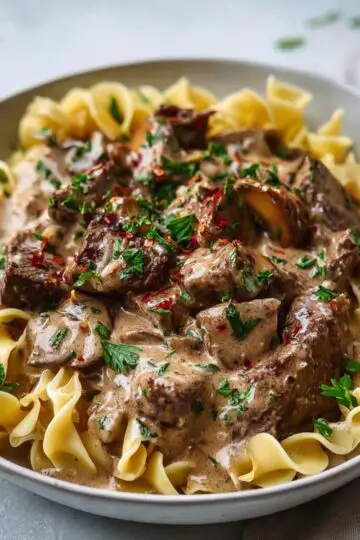 A hearty bowl of homemade beef stroganoff served over a bed of tender egg noodles. The dish features generous chunks of seared beef in a rich, creamy mushroom sauce, speckled with flecks of red pepper flakes and finely chopped fresh parsley. The sauce clings deliciously to the noodles and beef, giving a glossy, velvety finish. The noodles are cooked to a soft, slightly curled texture and sit nestled beneath the savory mixture. The dish is presented in a wide, off-white ceramic bowl on a light-colored surface, garnished with scattered parsley leaves for a fresh, rustic look.