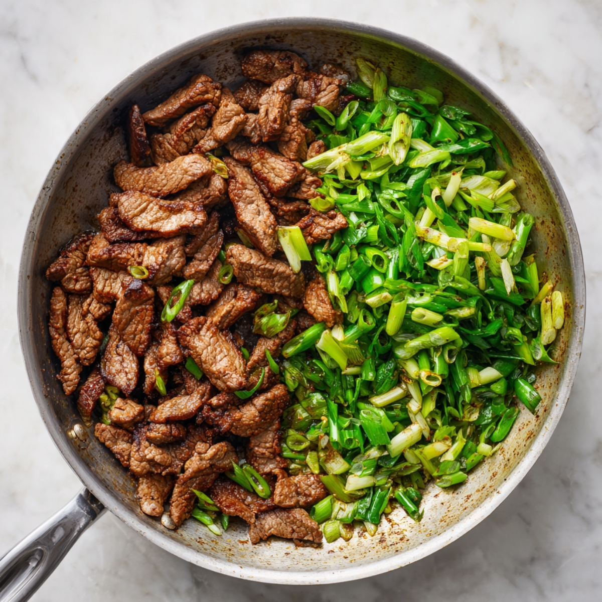A skillet on a marble countertop with browned beef strips on one side and fresh, chopped scallions on the other, both mid-sauté for a homemade meal.
