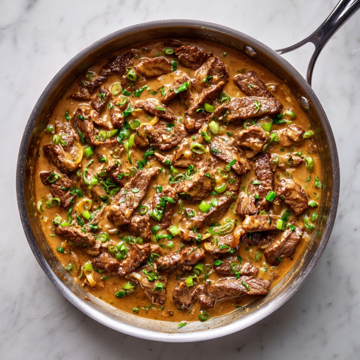 A skillet filled with creamy beef stroganoff, featuring tender beef strips in a rich brown sauce, topped with chopped green onions and parsley on a marble countertop.