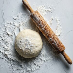 Ball of flour-dusted dough and a wooden rolling pin on a white countertop, surrounded by scattered flour in a cozy, homemade baking scene.