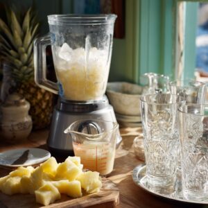 Blender filled with ice and pineapple, glasses on a tray, with measuring cup and cutting board in a sunny kitchen.