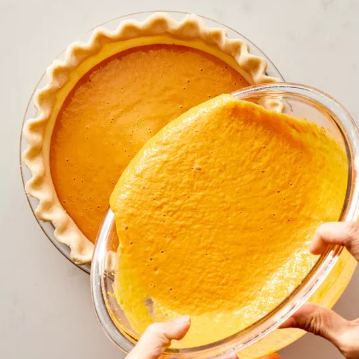 Homemade pumpkin pie filling being poured from a glass bowl into an unbaked pie crust with crimped edges, on a light kitchen counter in bright natural light.