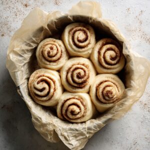 Unbaked cinnamon rolls in a parchment-lined round pan, tightly swirled with cinnamon-sugar and ready to bake, on a lightly dusted countertop.