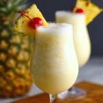 Homemade Pina Colada Recipe in a tall glass, topped with froth and garnished with a pineapple wedge and cherry, with a second drink and whole pineapple in the background.