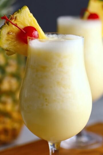 Homemade Pina Colada Recipe in a tall glass, topped with froth and garnished with a pineapple wedge and cherry, with a second drink and whole pineapple in the background.