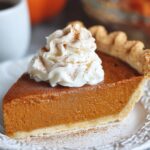 A slice of homemade pumpkin pie with a flaky crust, topped with whipped cream and cinnamon, on a white decorative plate with a coffee mug and warm autumn colors in the blurred background.