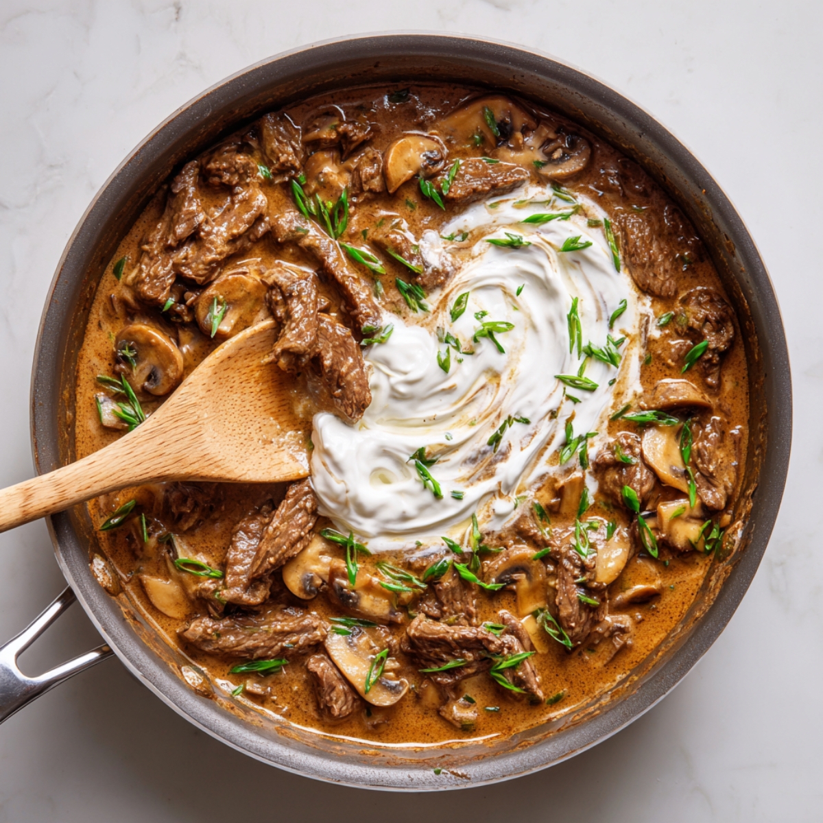 A skillet of homemade beef stroganoff being stirred with a wooden spoon, featuring tender beef strips, sliced mushrooms, a swirl of sour cream, and garnished with chopped green onions on a marble countertop.