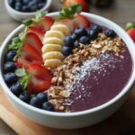 A bowl of Acai Bowl Recipe topped with sliced strawberries, bananas, blueberries, coconut shavings, and crushed nuts, arranged into neat sections.