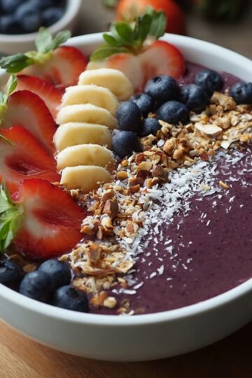 A bowl of Acai Bowl Recipe topped with sliced strawberries, bananas, blueberries, coconut shavings, and crushed nuts, arranged into neat sections.