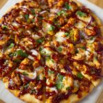 A freshly baked homemade BBQ chicken pizza sliced into pieces, topped with melted mozzarella cheese, tender chunks of BBQ chicken, thin slices of red onion, diced green bell peppers, fresh cilantro leaves, and drizzled with rich, glossy BBQ sauce. The crust is golden brown and slightly crispy. The pizza rests on parchment paper over a wooden cutting board, with natural lighting enhancing its warm, inviting, and rustic appearance.