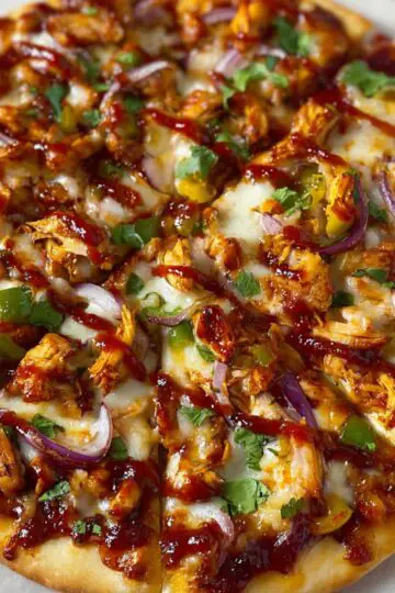 A freshly baked homemade BBQ chicken pizza sliced into pieces, topped with melted mozzarella cheese, tender chunks of BBQ chicken, thin slices of red onion, diced green bell peppers, fresh cilantro leaves, and drizzled with rich, glossy BBQ sauce. The crust is golden brown and slightly crispy. The pizza rests on parchment paper over a wooden cutting board, with natural lighting enhancing its warm, inviting, and rustic appearance.