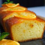 A close-up, side-angle shot of a homemade orange cake recipe sliced into thick pieces on a dark slate board. The front slice reveals a tender, fine-crumb interior and a glossy, amber-hued glaze dripping slightly down its golden-brown crust. Each slice is crowned with a thin, candied orange round sprinkled with bright green zest. In the foreground, a fresh mint sprig rests next to an extra orange wheel, all bathed in soft, natural light that casts gentle shadows and gives the scene a warm, cozy kitchen feel.