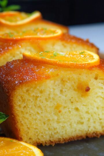 A close-up, side-angle shot of a homemade orange cake recipe sliced into thick pieces on a dark slate board. The front slice reveals a tender, fine-crumb interior and a glossy, amber-hued glaze dripping slightly down its golden-brown crust. Each slice is crowned with a thin, candied orange round sprinkled with bright green zest. In the foreground, a fresh mint sprig rests next to an extra orange wheel, all bathed in soft, natural light that casts gentle shadows and gives the scene a warm, cozy kitchen feel.