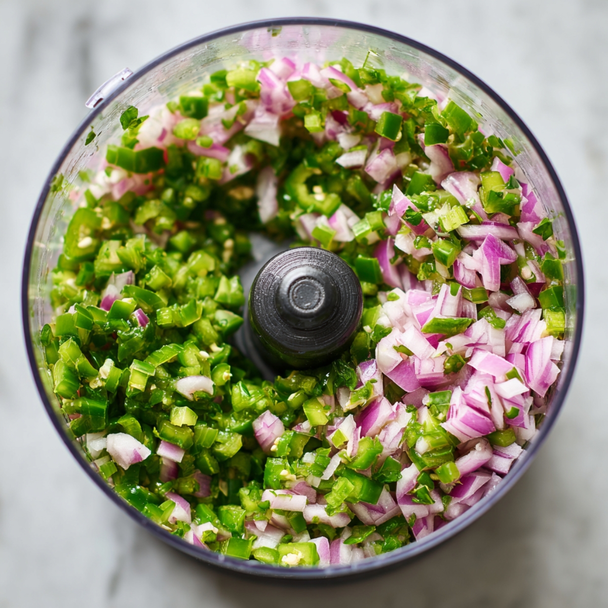 Close-up overhead of a food-processor bowl containing finely diced green jalapeños and red onion around the blade, on a marble surface.