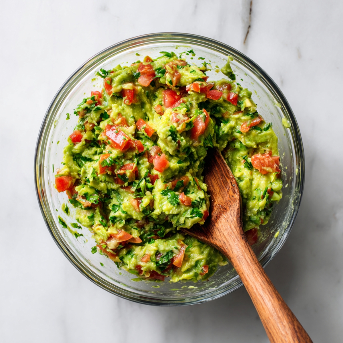 Top-down view of chunky avocado-tomato-cilantro guacamole in a clear glass bowl, with a wooden spoon coated in guacamole resting inside.