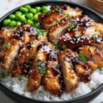 A black ceramic bowl of steamed white rice topped with sliced, glossy teriyaki chicken sprinkled with sesame seeds and herbs, a cluster of bright edamame at the rim, and a small wooden bowl of extra sauce on a white marble surface under soft natural light.