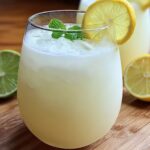 A glass of creamy Brazilian lemonade with ice, garnished with a lemon slice and mint, on a wooden surface with lemon and lime slices in the background.