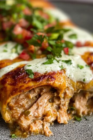 Three homemade chicken enchiladas in a dark baking dish, smothered in red sauce and melted cheese, topped with a dollop of sour cream, cilantro, and diced red onion.