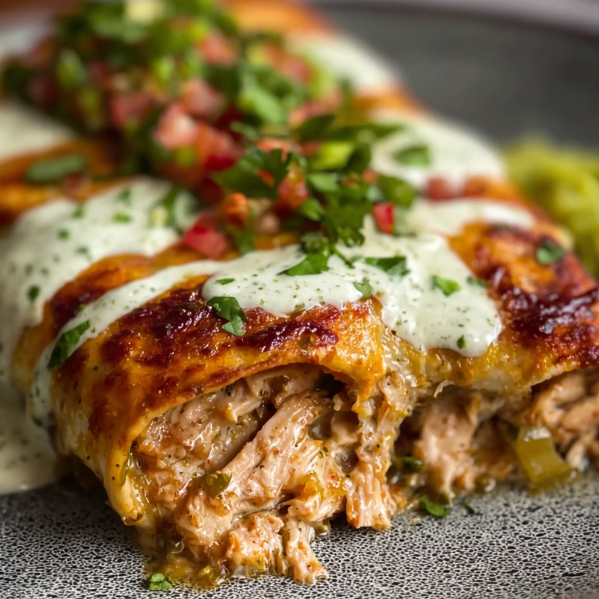 Three homemade chicken enchiladas in a dark baking dish, smothered in red sauce and melted cheese, topped with a dollop of sour cream, cilantro, and diced red onion.