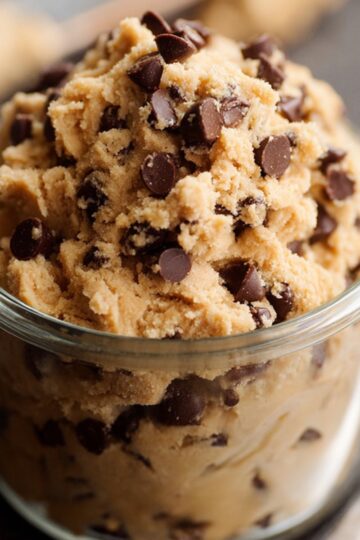 An overhead view of three generous scoops of homemade cookie dough ice cream nestled in a simple white bowl. The off-white, creamy base is studded with irregular dark chocolate cookie bits. A vintage silver spoon rests against the side, and a few extra cookie crumbs are scattered on the gray stone surface around the bowl.