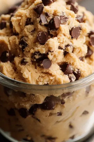 An overhead view of three generous scoops of homemade cookie dough ice cream nestled in a simple white bowl. The off-white, creamy base is studded with irregular dark chocolate cookie bits. A vintage silver spoon rests against the side, and a few extra cookie crumbs are scattered on the gray stone surface around the bowl.