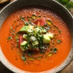 A bowl of homemade gazpacho with a smooth red soup topped with diced cucumber, tomatoes, and fresh cilantro. Olive oil drizzles on top with freshly ground black pepper. The rustic, speckled bowl adds a cozy, homemade feel.