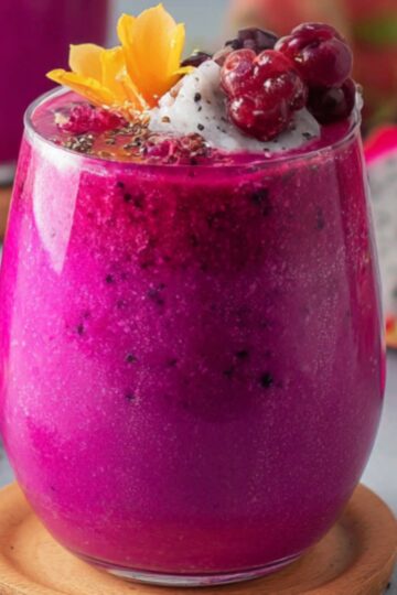 A vibrant pink dragon fruit smoothie served in a glass, topped with coconut flakes, berries, and a yellow flower, with dragon fruit slices in the background.