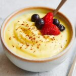 A white ceramic bowl filled with creamy yellow Custard Recipe, topped with fresh strawberries and blueberries and a light dusting of nutmeg, with a spoon resting in the custard.