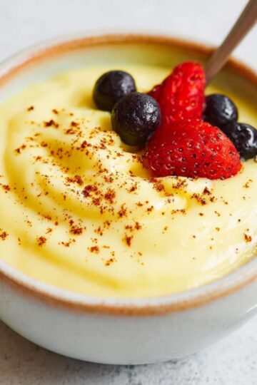 A white ceramic bowl filled with creamy yellow Custard Recipe, topped with fresh strawberries and blueberries and a light dusting of nutmeg, with a spoon resting in the custard.