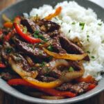 Vue rapprochée d’une assiette creuse en grès gris : un généreux lit de riz blanc moelleux sur la moitié gauche, surmonté à droite d’un mélange de fines tranches de bœuf tendre,pepper steak recipe de lamelles de poivron et d’oignon légèrement caramélisées, agrémentées de brins de ciboule ciselée et d’une sauce brune brillante.