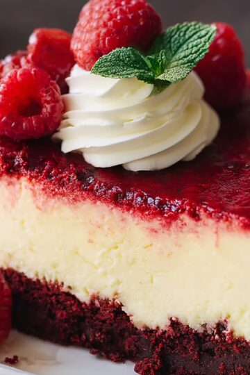 Close-up of a cheesecake slice with red velvet crust, creamy center, raspberry topping, whipped cream rosette, and fresh raspberries on the side.