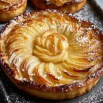 A close-up of a perfectly arranged tarte aux pommes in a tart pan, with thin, overlapping apple slices in concentric circles, glazed to a golden shine and lightly dusted with powdered sugar.