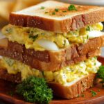 “Three-layer toasted bread Egg Sandwich filled with fried eggs and melted cheese, a runny yolk dripping down the side, garnished with sprigs of fresh thyme on a pale tabletop.”