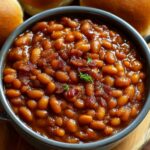 A rustic black pot on a wood round, brimming with thick, caramelized baked beans recipe studded with bacon bits and garnished with fresh parsley, surrounded by dinner rolls.