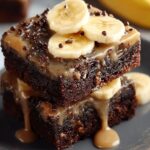 Two thick Banana Brownies stacked on a dark plate, topped with fresh banana slices and a glossy caramel drizzle, with a whole banana blurred in the background.