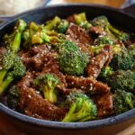 Close-up skillet of Beef and Broccoli Recipe in glossy brown sauce, sprinkled with sesame seeds, rice blurred in background