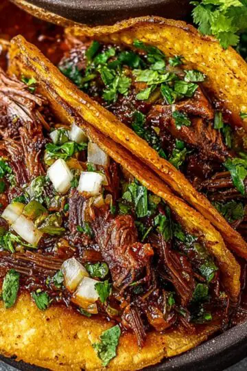 Two crispy birria tacos filled with juicy, shredded beef and topped with fresh cilantro and diced onions, served with a bowl of rich consommé and lime wedges on a dark rustic plate.