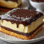 Close‑up of a square slice of Boston cream pie showing a golden sponge cake base, thick vanilla custard layer, and glossy chocolate ganache dripping over the edges, topped with white chocolate shavings on parchment