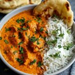 A white bowl half‑filled with fluffy basmati rice drizzled with cream, the other half with orange‑red Butter Chicken Recipe pieces in rich tomato‑cream sauce, garnished with chopped cilantro, served with a folded naan on the side on a dark stone table.